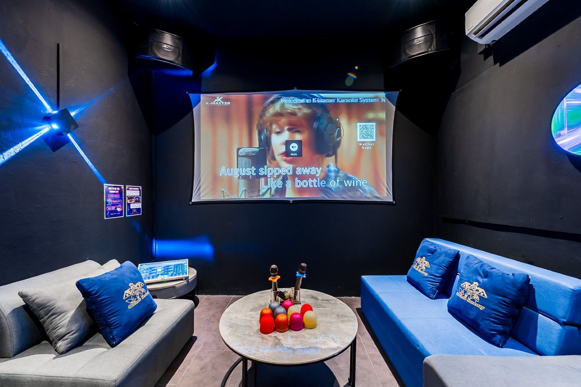 A karaoke room with a large screen, two couches, a table with microphones, and a wall with a QR code.