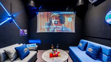 A karaoke room with a large screen, two couches, a table with microphones, and a wall with a QR code.