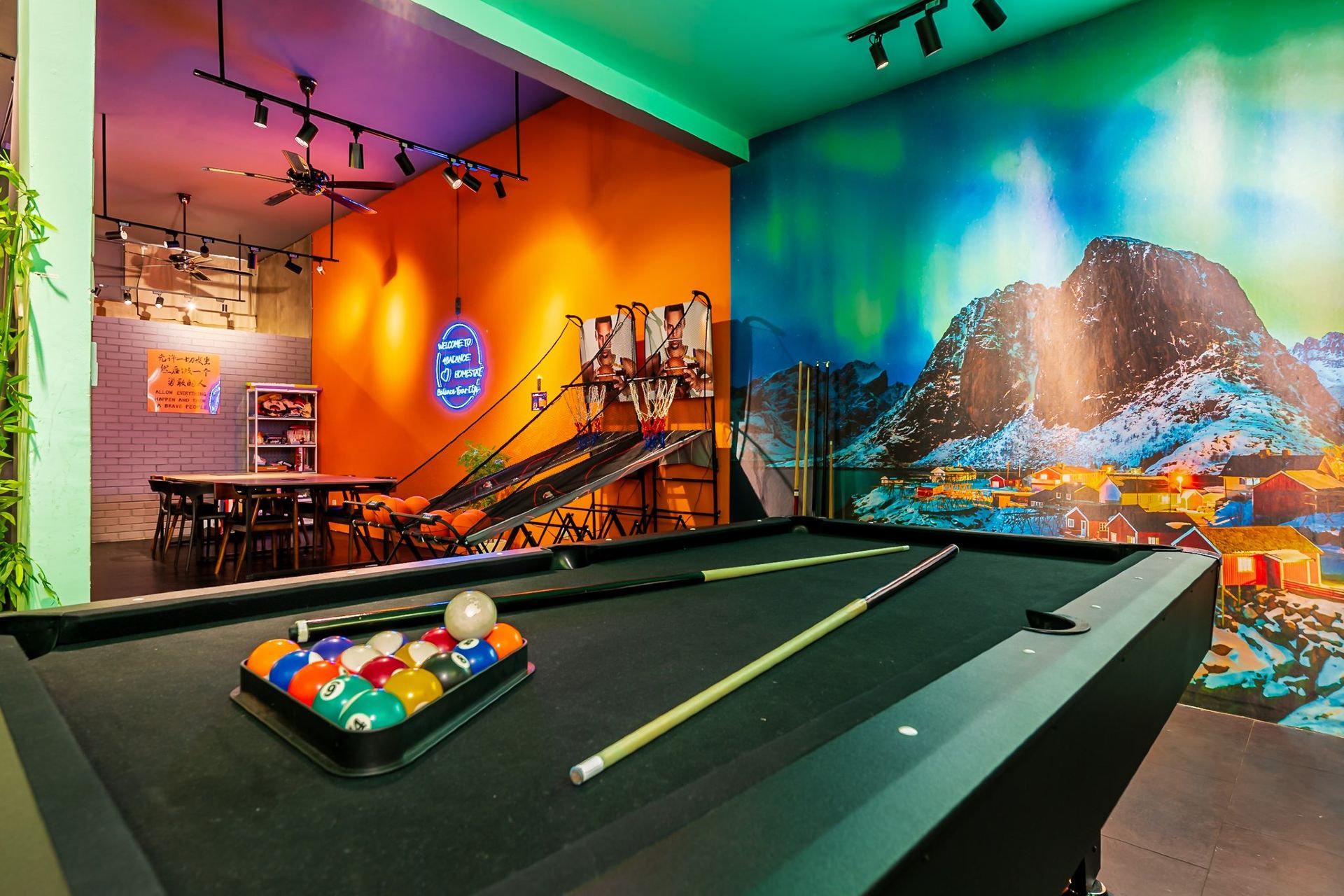 Una mesa de billar con bolas y tacos, un letrero de neón, un mural de una montaña nevada y una zona de bar con taburetes y una estantería.