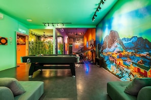 A billiards room with a pool table, green sofas, a mural, and a staircase.