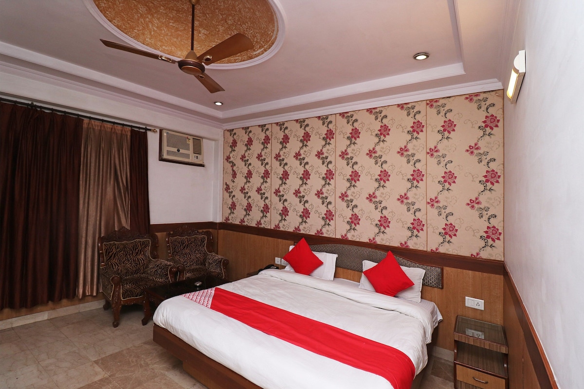 A hotel room with a bed, a ceiling fan, a window with curtains, a wooden headboard, and a floral wallpaper.