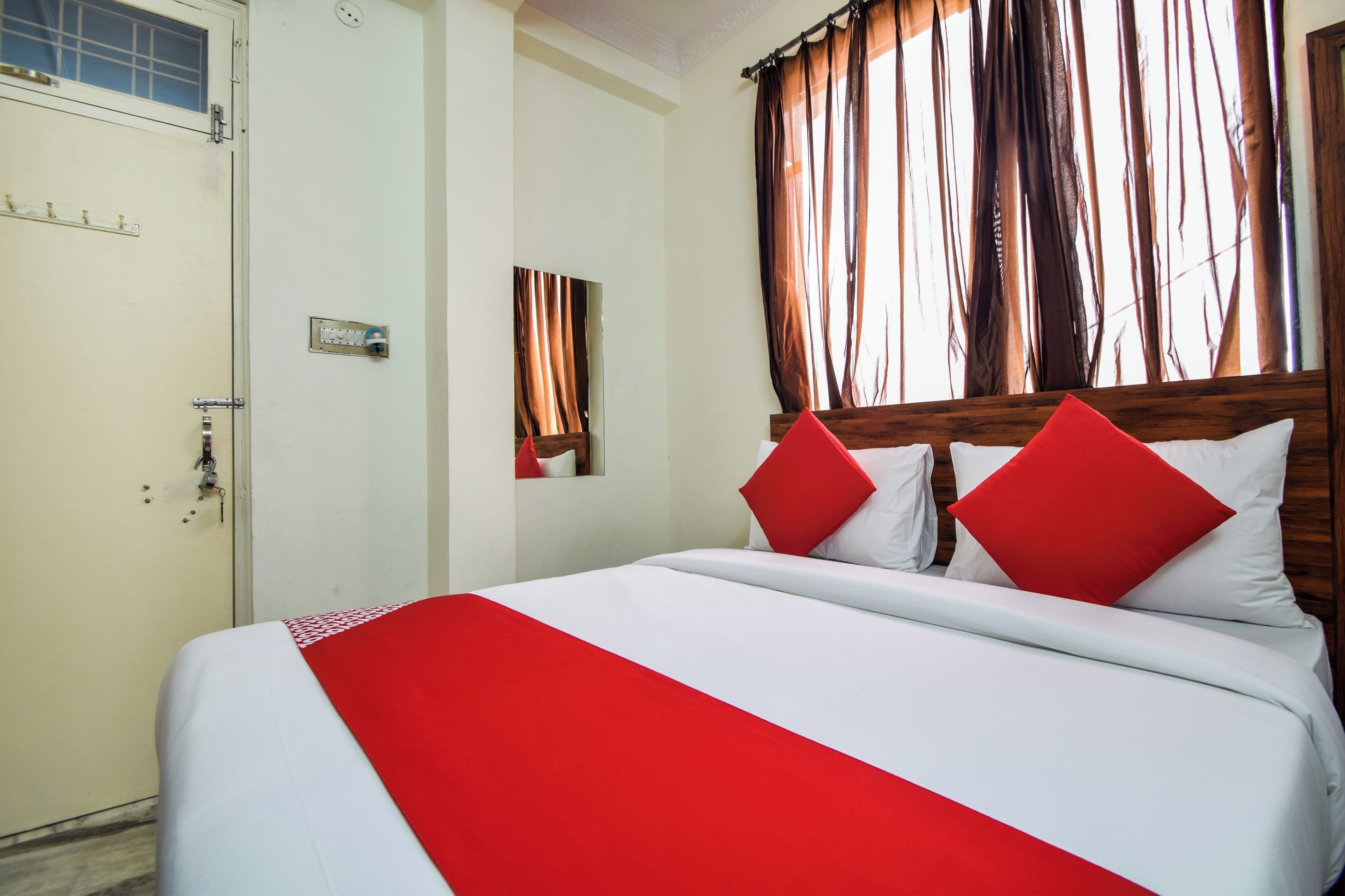 A hotel room with a bed, a window with curtains, a door, and a wall-mounted light switch.