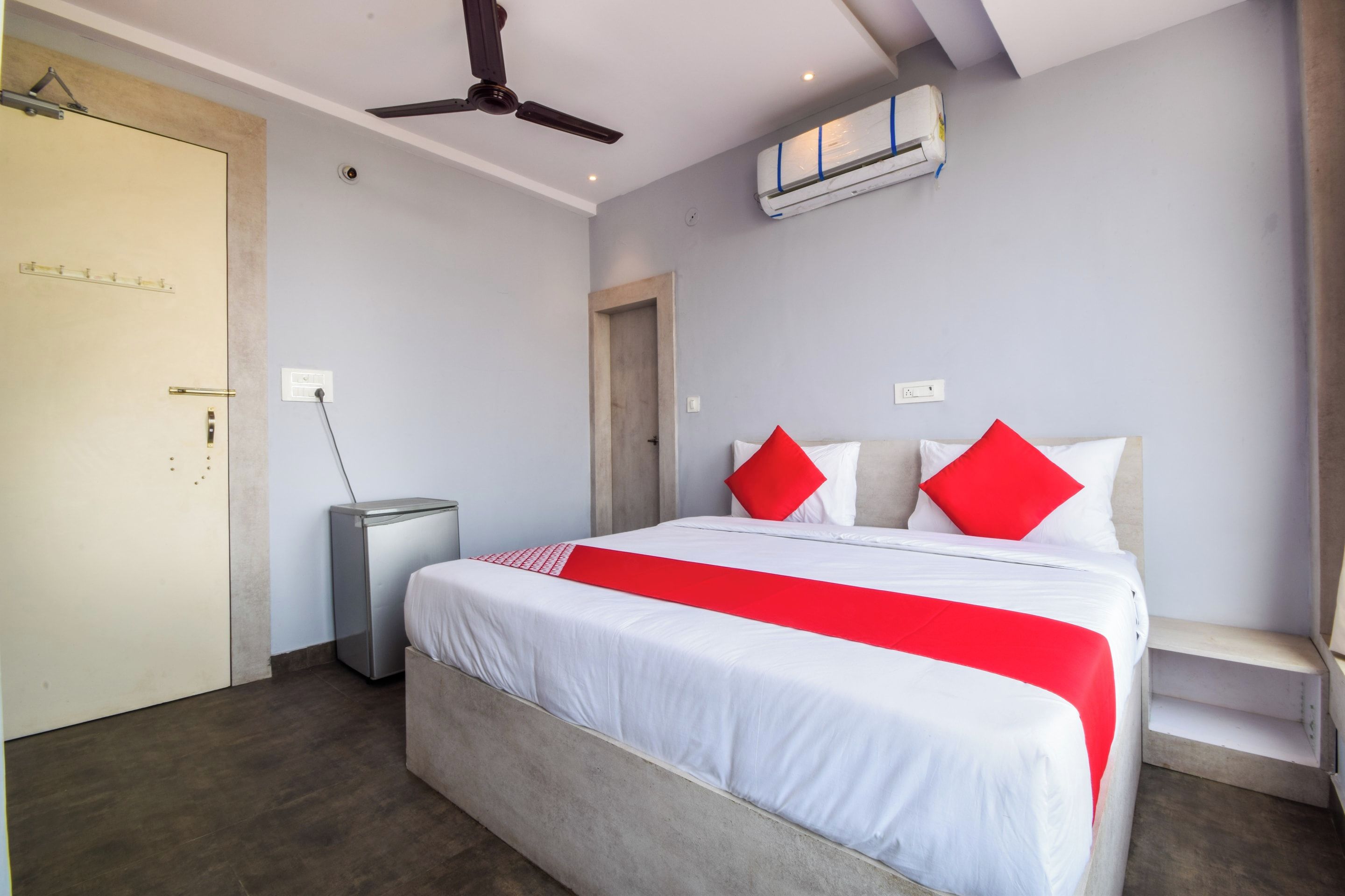 Una habitación de hotel con una cama, un ventilador de techo, un equipo de aire acondicionado, una mesita de noche y una puerta.
