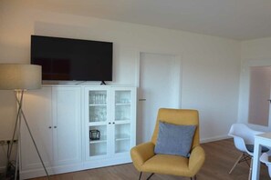 A room with a television on a cabinet, a yellow armchair with a pillow, and a white table with chairs. - Holiday apartment Friesenperle (Horumersiel)