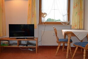 A room with a television on a wooden stand, a dining table with chairs, and a window with curtains. - KLA8 in the Säuling House (Schwangau)