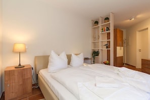 Ein ordentlich bezogenes Bett mit weißen Leinen und Kissen, eine Nachttischlampe, ein Holennachttisch und ein Bücherregal mit verschiedenen Gegenständen.