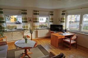 A living room with a wooden desk, a television, a round table with a vase of flowers, and a view of the street through large windows. - Holiday apartment with 1 bedroom (Ahlbeck)