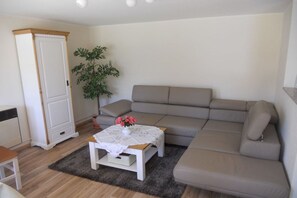 A living room with a beige sofa, a white table with a vase of flowers, a tall white cabinet, and a potted plant. - Holiday home with 1 bedroom (Schwielochsee)