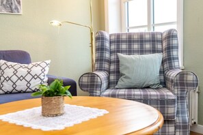 Ein Zimmer mit Sesseln im Karomuster, einem blauen Sofa, einem Holztisch mit Pflanze und einer Stehlampe.