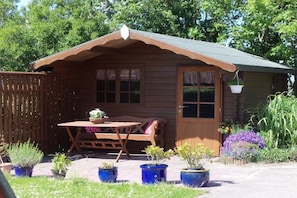 Eine Holzhütte mit Veranda, Tisch und Stühlen. Kübelpflanzen und ein Holzzaun sind ebenfalls vorhanden.