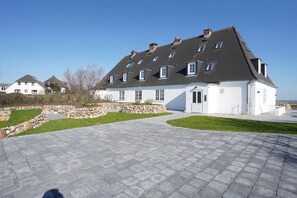 Een groot wit gebouw met een grijs dak, meerdere ramen en een verharde oprit.