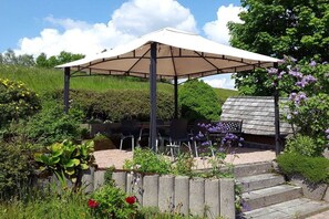 A covered outdoor seating area with a table and chairs, surrounded by a garden with various plants and flowers. - Holiday home with 2 bedrooms (St. Georgen\/ Langenschiltach)