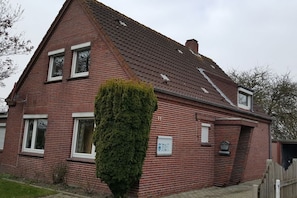 A red brick house with a sloped roof, a small front yard, and a tall green hedge. - Holiday apartment with 1 bedroom (Norden)