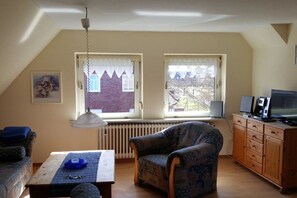 A room with a sloped ceiling, two windows with curtains, a radiator, a wooden cabinet with a television, and a patterned armchair. - Holiday apartment with 1 bedroom (Norden)