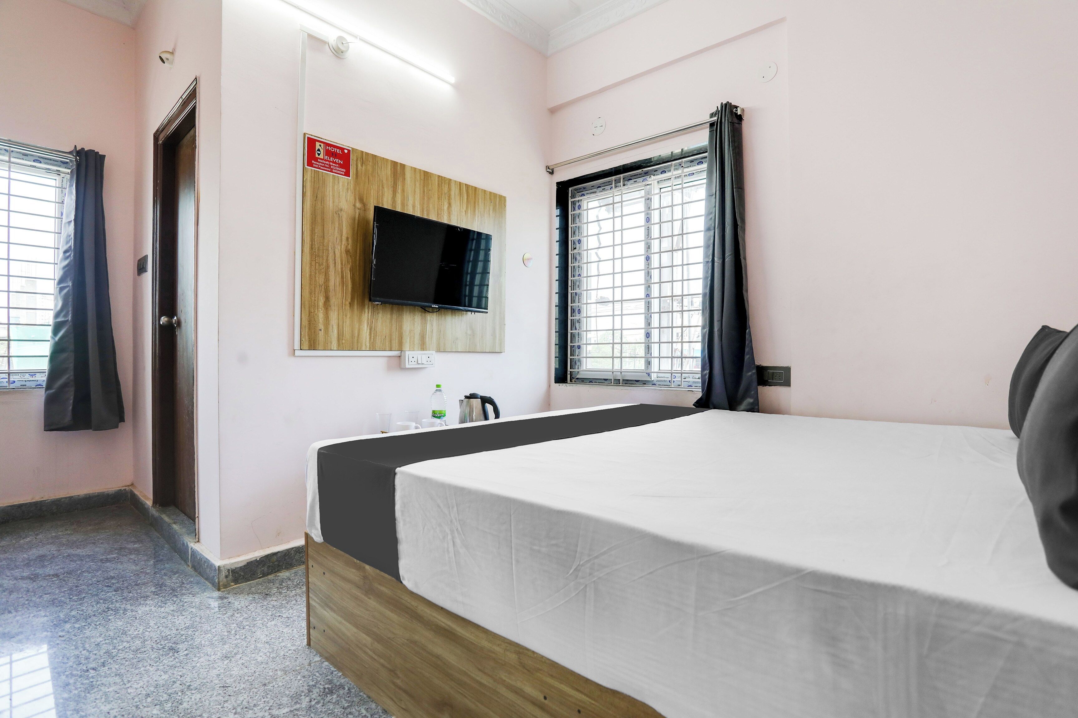 A hotel room with a bed, a television mounted on a wooden panel, a window with curtains, and a small table with a kettle and cups.