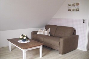 A room with a brown sofa, a wooden coffee table with a lace doily, and a wall-mounted picture frame. - Holiday apartment in Föhr with 1 bedroom (Oldsum)