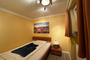 A bedroom with a bed, a nightstand, a lamp, a painting on the wall, and a window with curtains. - Holiday apartment with 2 bedrooms (Westerland)