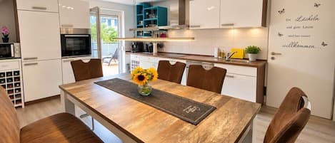 A modern kitchen with white cabinets, a wooden dining table, and a welcome sign.