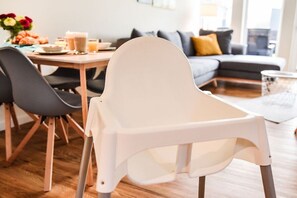 Ein Essbereich mit einem weißen Hochstuhl, einem Tisch mit Blumenvase und einer Couch im Hintergrund.