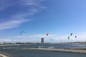 Eine Küstenlandschaft mit mehreren Kitesurfern, einem Gewässer und einem Gebäude in der Ferne.