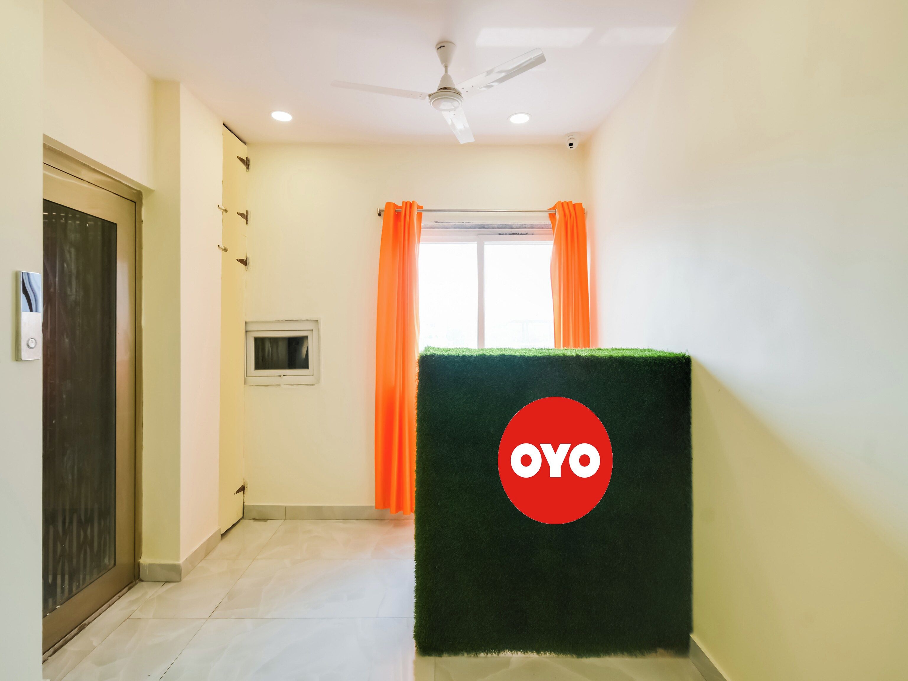 A hotel corridor with a ceiling fan, a window with orange curtains, and a green artificial turf with a logo.