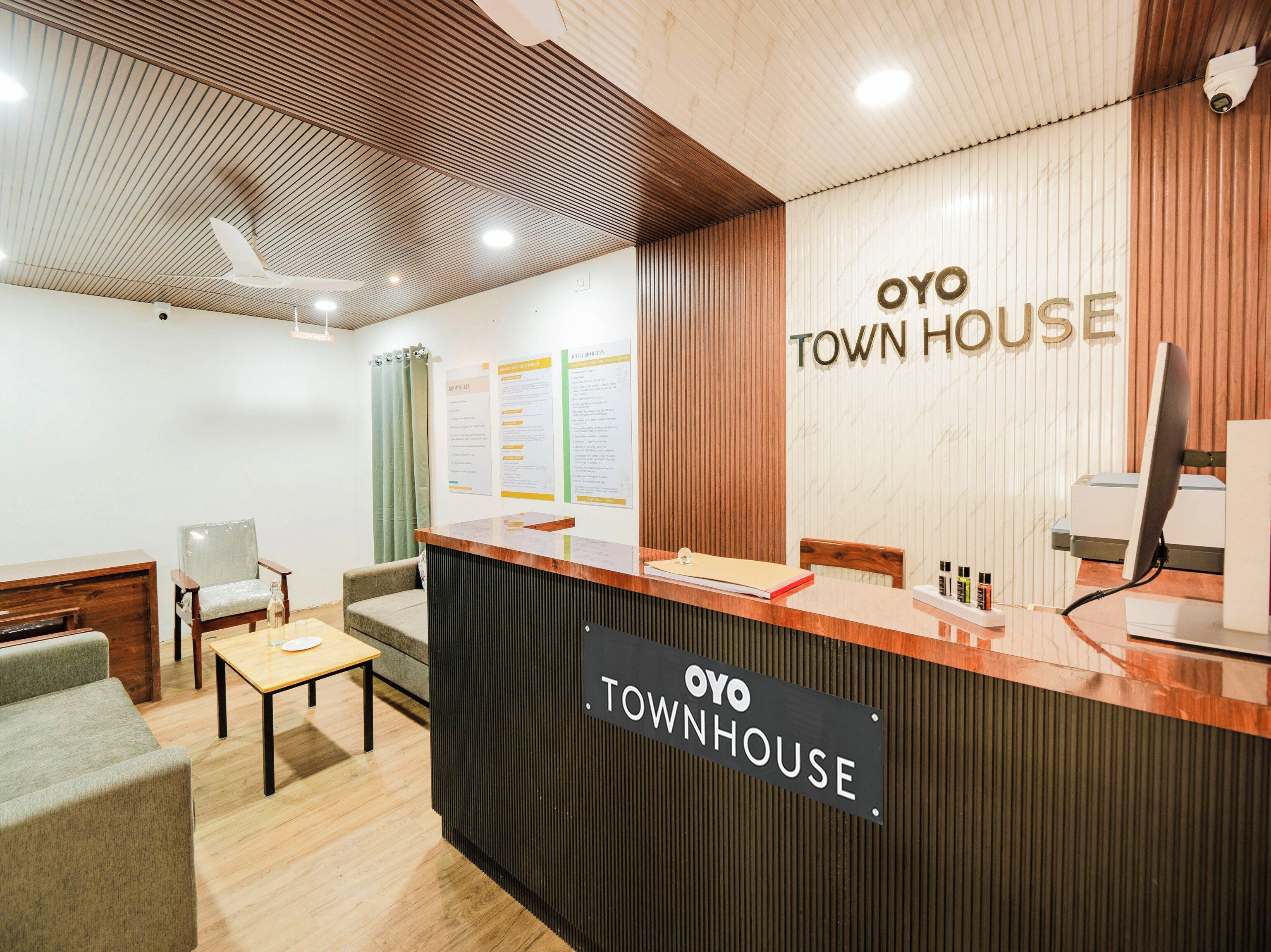 Una recepción con un cartel que dice 'OYO TOWN HOUSE' y un monitor de computadora. Se observan sillas y una mesa en primer plano.