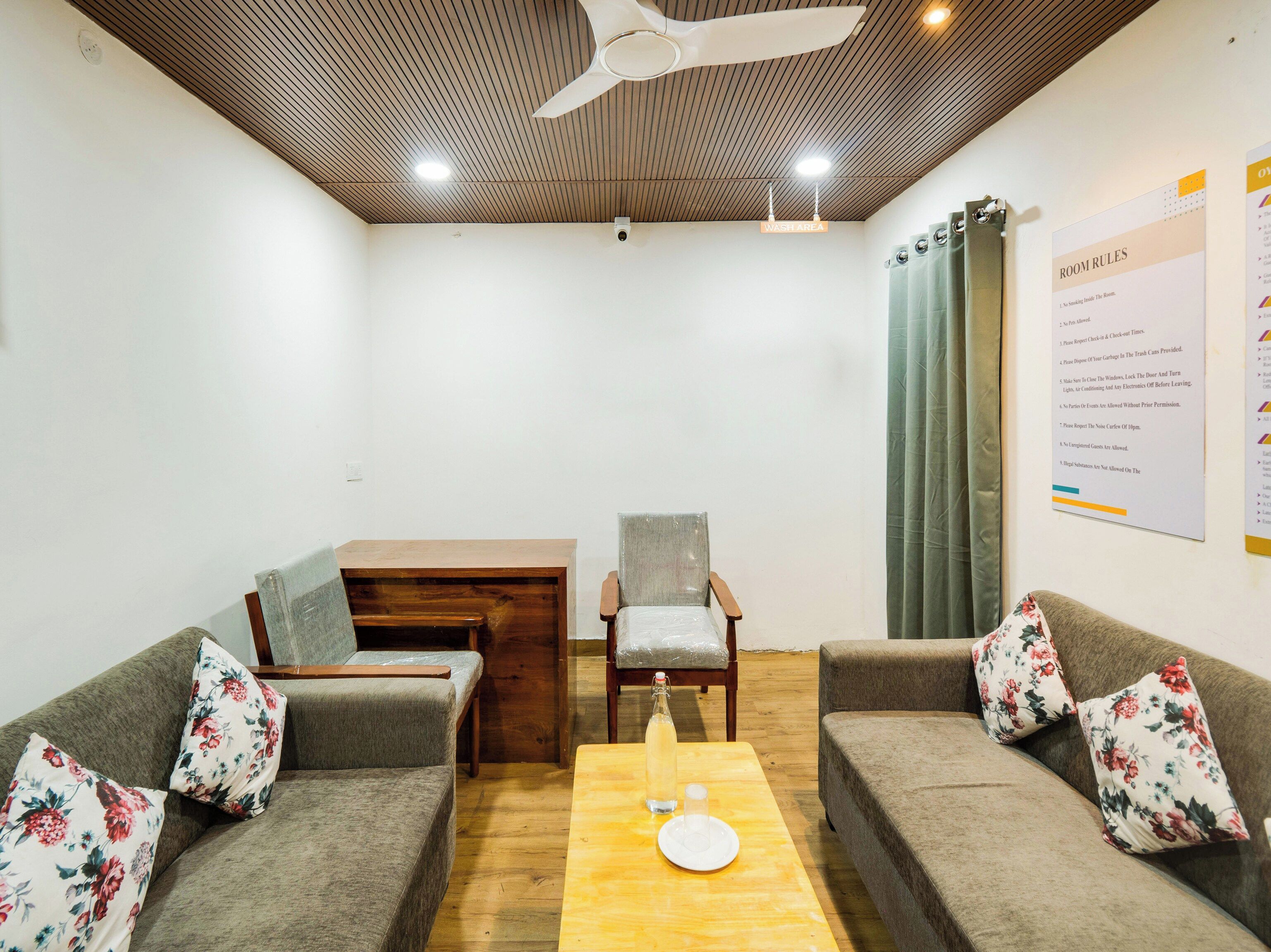 Una habitación con dos sofás, una mesa de madera, una silla, un ventilador de techo y una pared con las normas de la habitación.