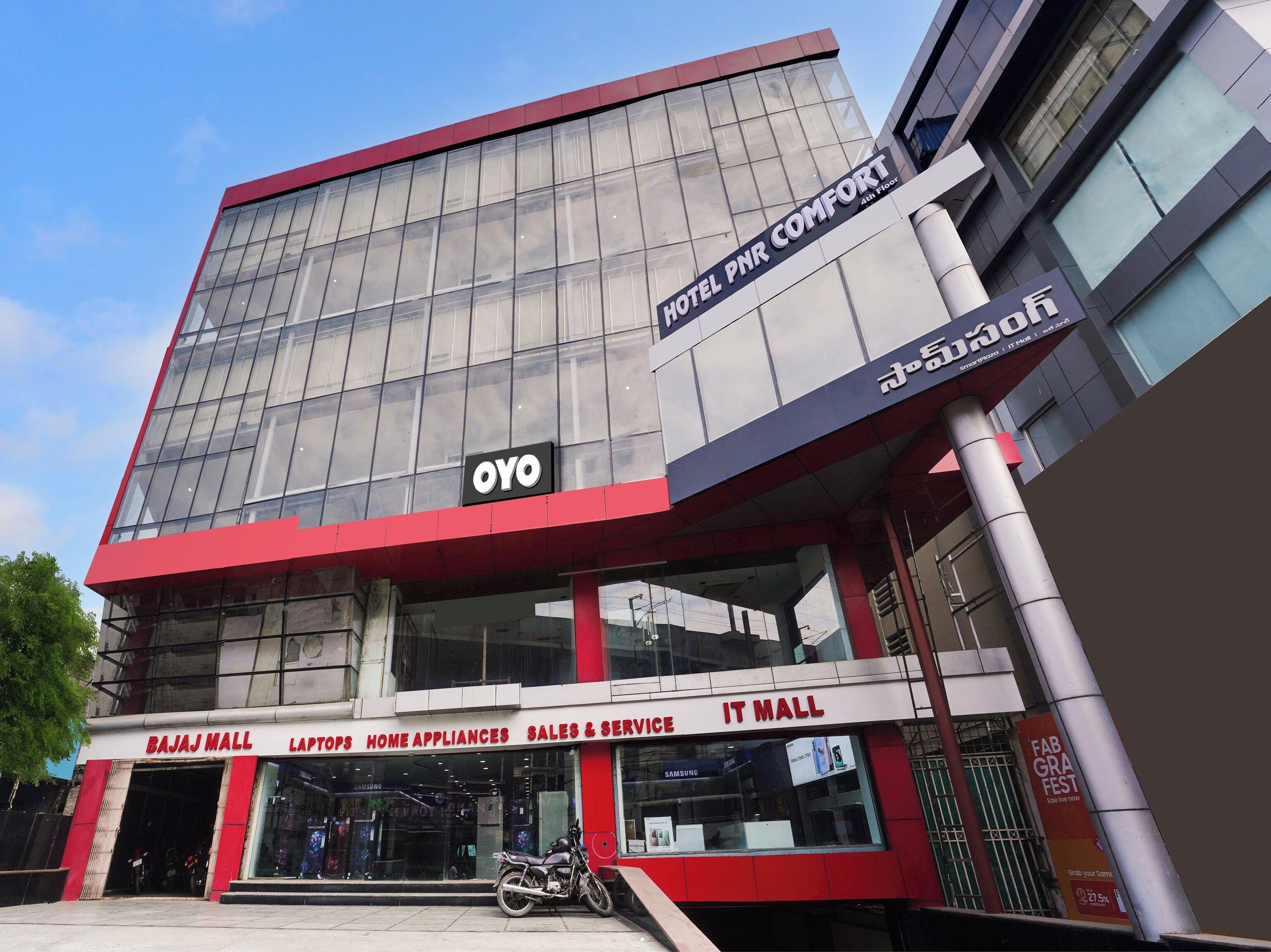 A modern building with a glass facade, a sign for 'Hotel PNR Comfort' on the 4th floor, and a 'Bajaj Mall' with various retail outlets.