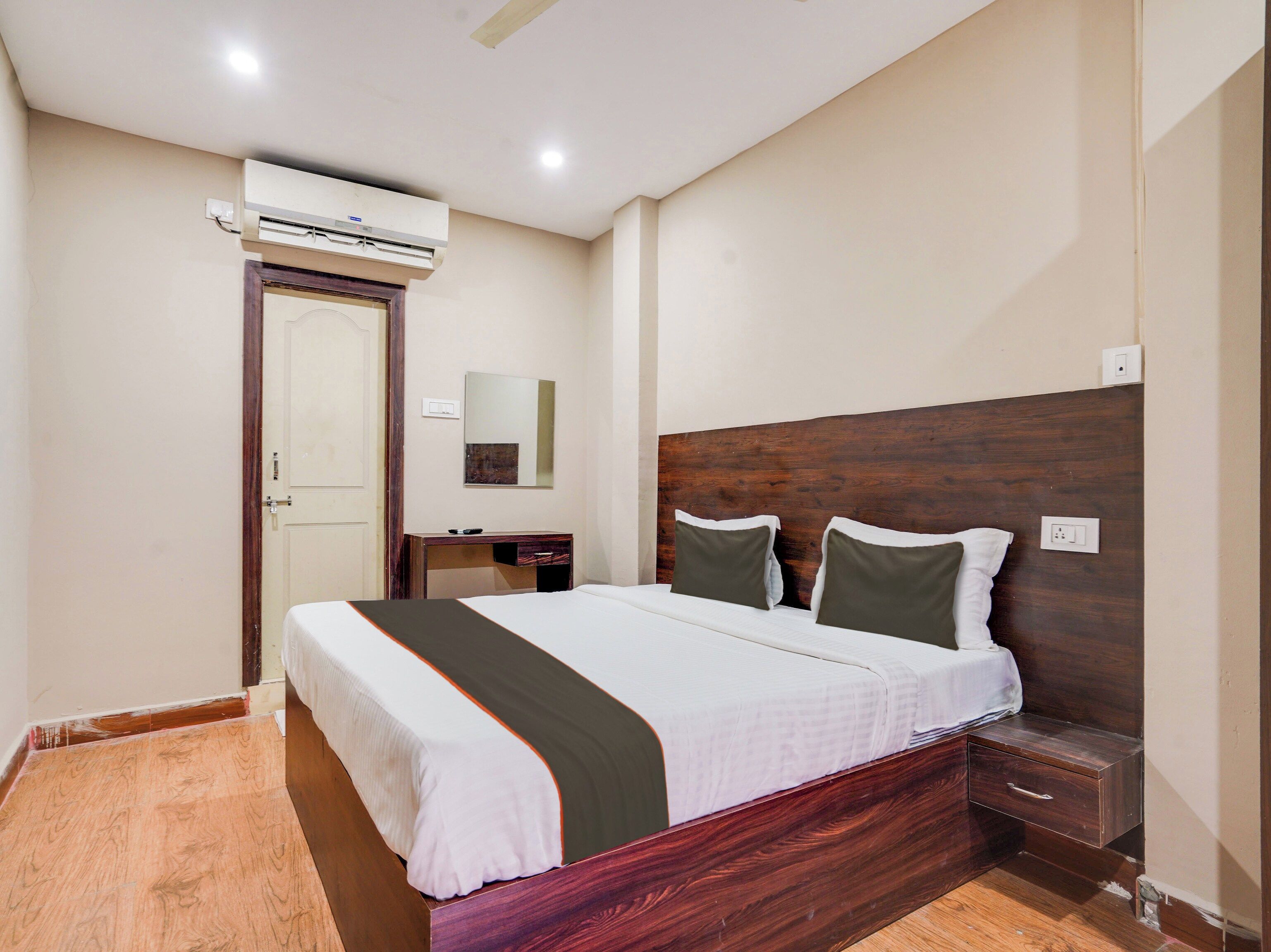 A hotel room with a bed, bedside table, nightstand, and an air conditioning unit.