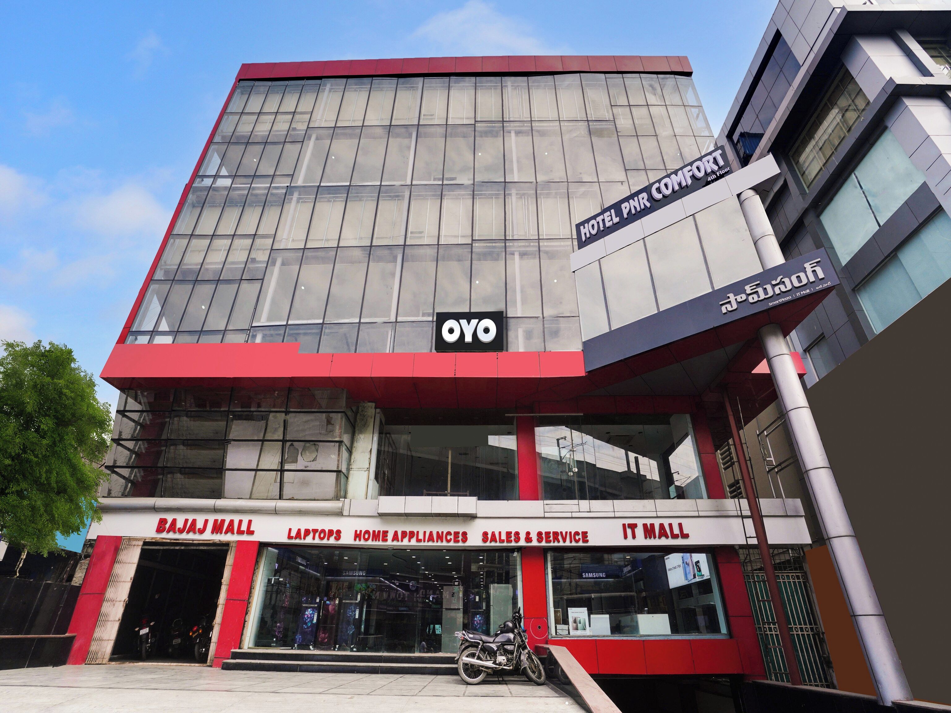 A modern building with a glass facade, a sign for 'Hotel PNR Comfort' on the upper right, and 'Bajaj Mall' with 'Laptops, Home Appliances, Sales & Service, IT Mall' on the lower left.