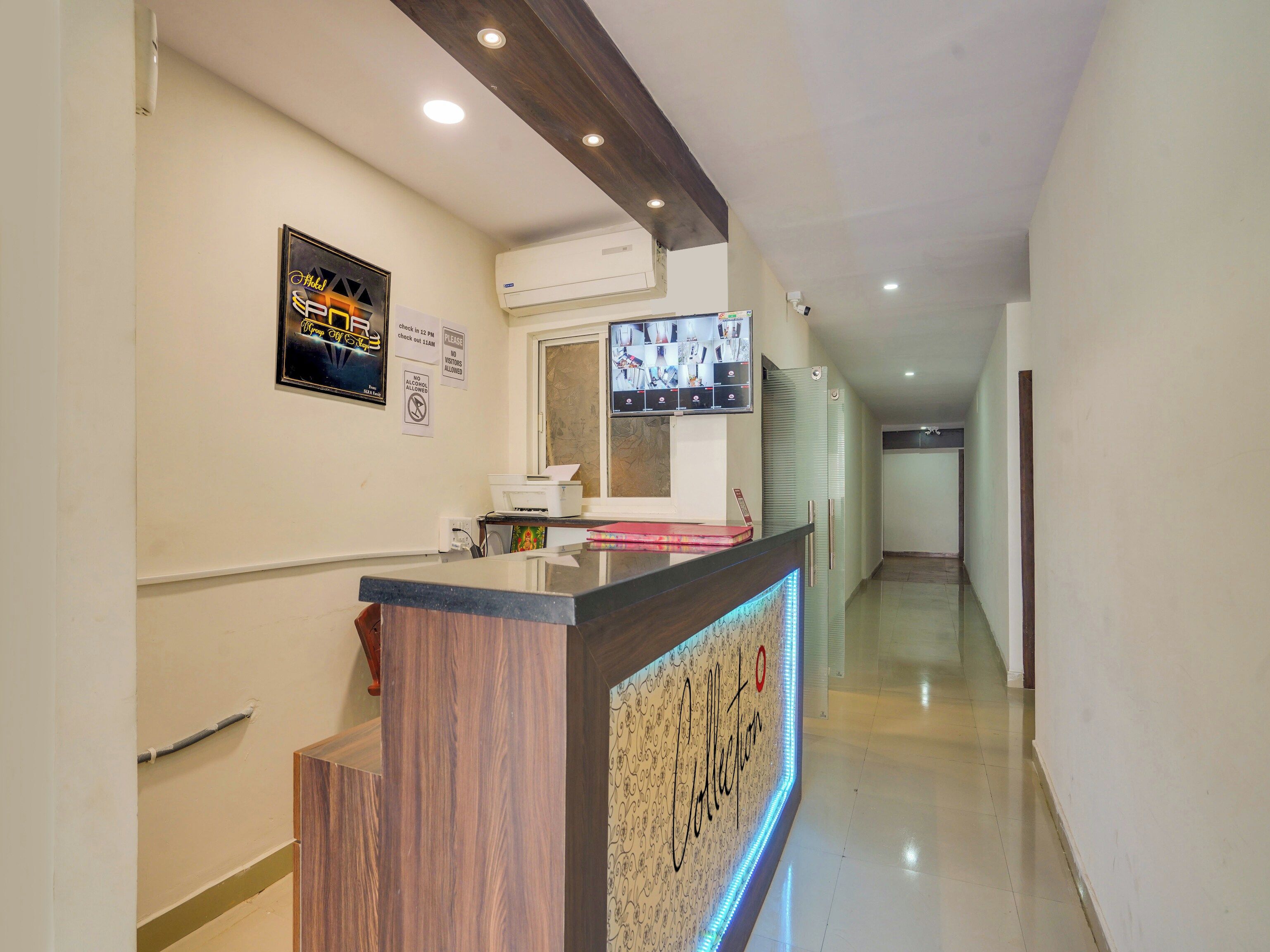 A hotel reception area with a wooden reception desk, a digital screen displaying images, and a wall-mounted air conditioning unit.