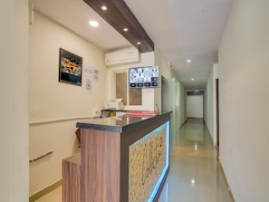 A hotel reception area with a wooden reception desk, a digital screen displaying images, and a wall-mounted air conditioning unit. - Super Collection O PNR COMFORT (Hyderabad)