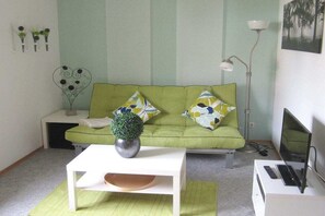 A living room with a green sofa, a white coffee table, a floor lamp, and a TV stand with a monitor. - Holiday apartment with 1 bedroom (Burrweiler)