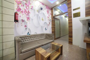 Un área de recepción con un sofá, una mesa de centro de vidrio, una pared decorada con motivos florales y mariposas, y un mostrador de recepción.
