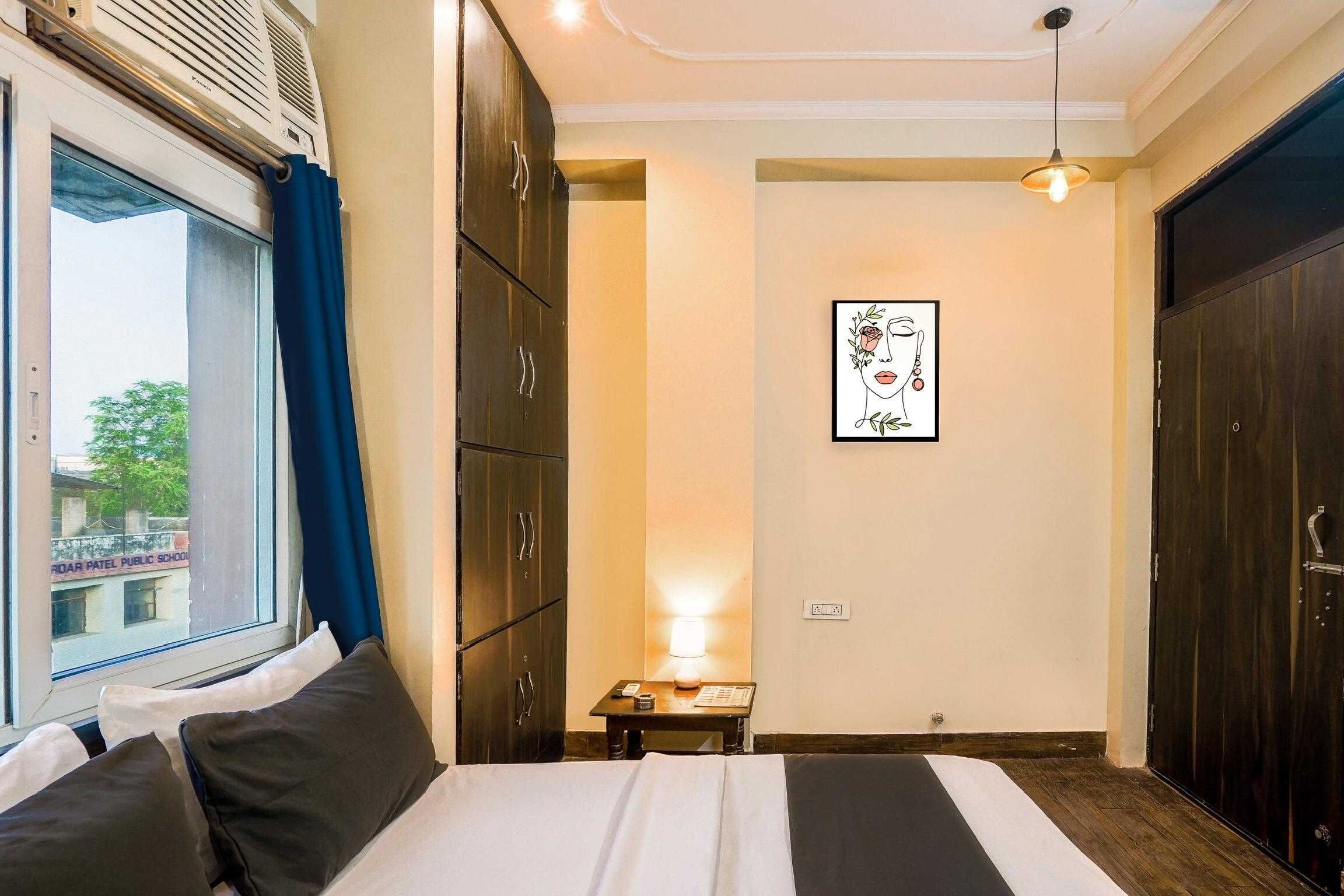 A hotel room with a bed, a bedside table, a lamp, a framed artwork, and a window with curtains.