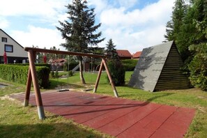 A playground with swings and a red rubberized surface, surrounded by greenery and a building with a sloped roof. - Hasselfelde House 24 (Hasselfelde)