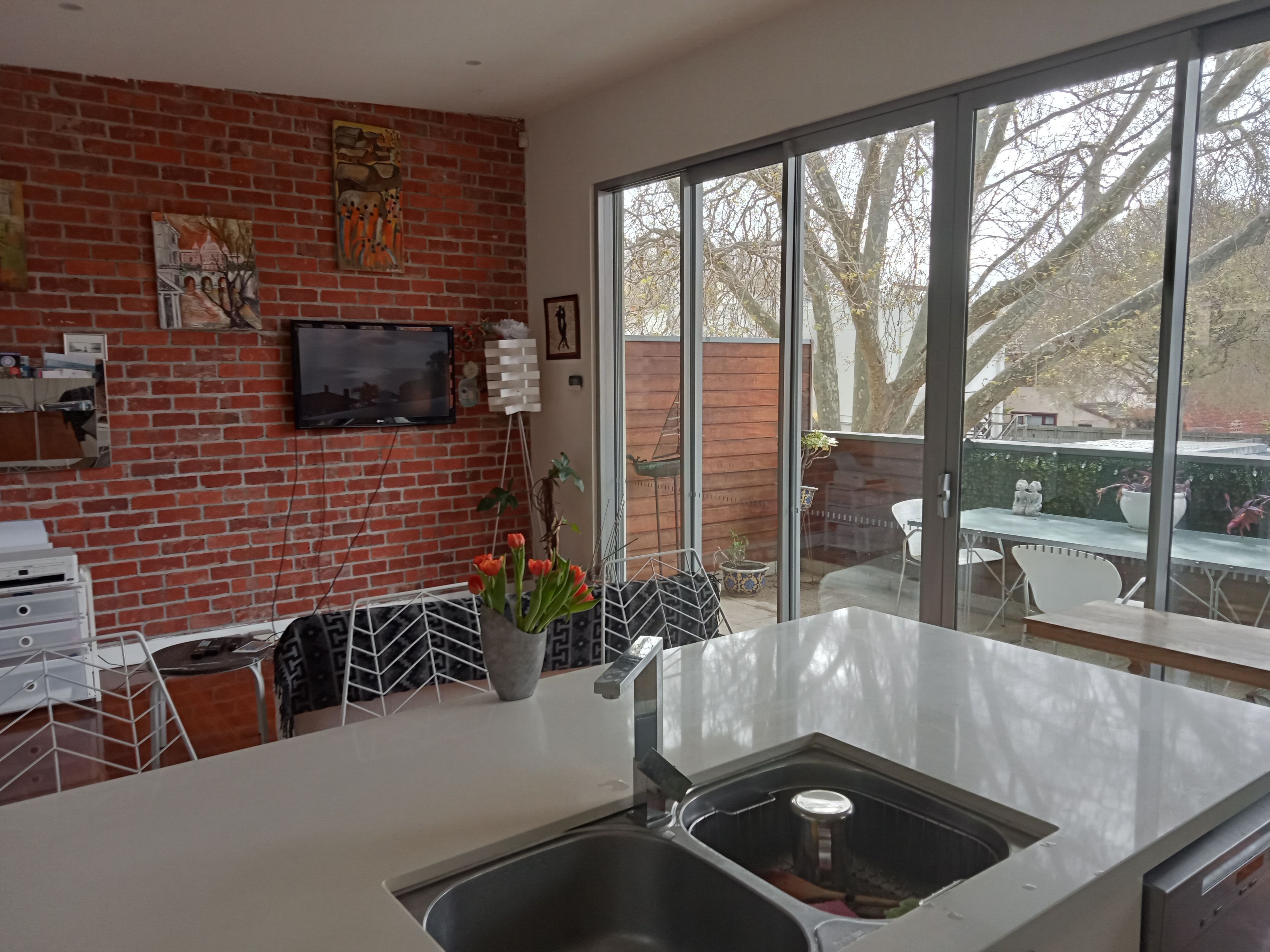 A kitchen with a brick wall, a TV mounted above the counter, and a large window offering a view of the outdoors.
