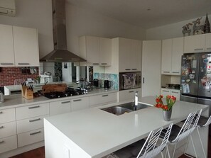 A modern kitchen with white cabinetry, a central island with bar stools, a stainless steel refrigerator, and a gas stove. -  A WONDERFUL TOWNHOUSE THAT IS ONE MINUTE FROM THE BEACH AND CENTRAL TO SHOPS. (Elwood)