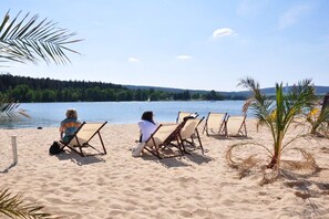A beach with two people seated on beach chairs facing a lake, surrounded by palm trees and a clear sky. - Holiday apartment in Erfurt with barbecue (Erfurt)