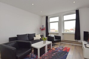 A modern living room with a black sofa, a white coffee table, and a colorful rug. There are two windows with black curtains, and a television on a stand. - Holiday apartment in Erfurt with barbecue (Erfurt)