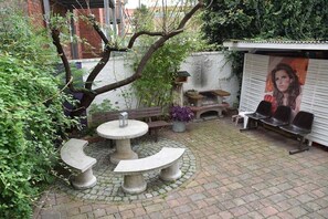 Een binnentuin met een stenen tafel en banken, omgeven door groen en een muur met een muurschildering.