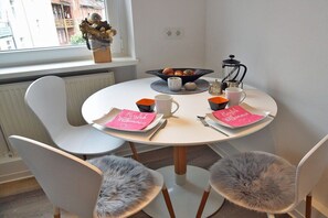 A round dining table with a white tablecloth, four chairs with fluffy seat cushions, a bowl of fruit, a coffee pot, and a vase with dried flowers on the windowsill. - Holiday apartment in Erfurt with barbecue (Erfurt)