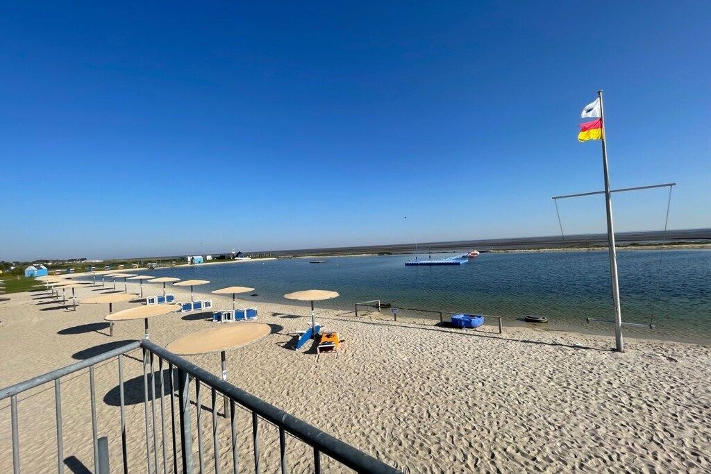 Une plage avec des parasols et des chaises longues, un mât avec un drapeau, et une étendue d'eau en arrière-plan.