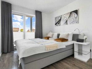 A modern bedroom with a large bed, a bedside table with a lamp, and a view of the outdoors through a sliding door. - Holiday Apartment With 2 Bedrooms (Kappeln)