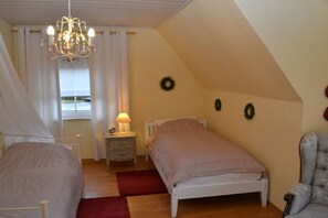 A bedroom with two beds, a chandelier, a nightstand with a lamp, and a view of the outdoors through a window. - Holiday home with 2 bedrooms (Papenburg)