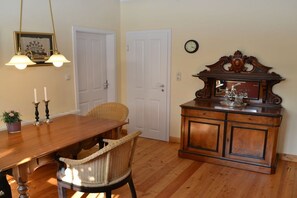 A dining area with a wooden table, wicker chairs, a cabinet with a clock, and a framed picture on the wall. - Holiday home with 2 bedrooms (Papenburg)