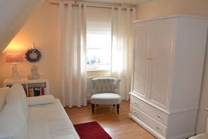 A room with a white sofa, a white wardrobe, a red rug, a chair, a lamp, and a window with curtains. - Holiday home with 2 bedrooms (Papenburg)