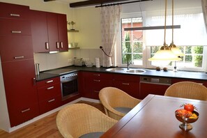 A kitchen with red cabinets, a wooden table, wicker chairs, and a window with curtains. - Holiday home with 2 bedrooms (Papenburg)