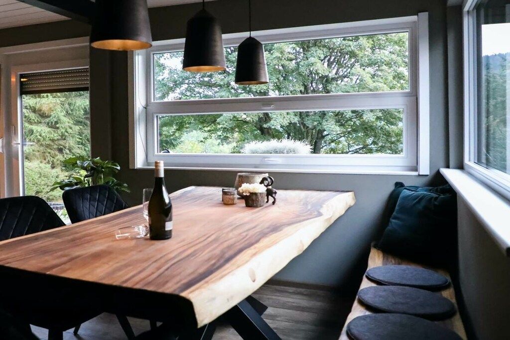 Uma área de refeições com mesa de madeira, cadeiras pretas, um banco com almofadas e luminárias pendentes.