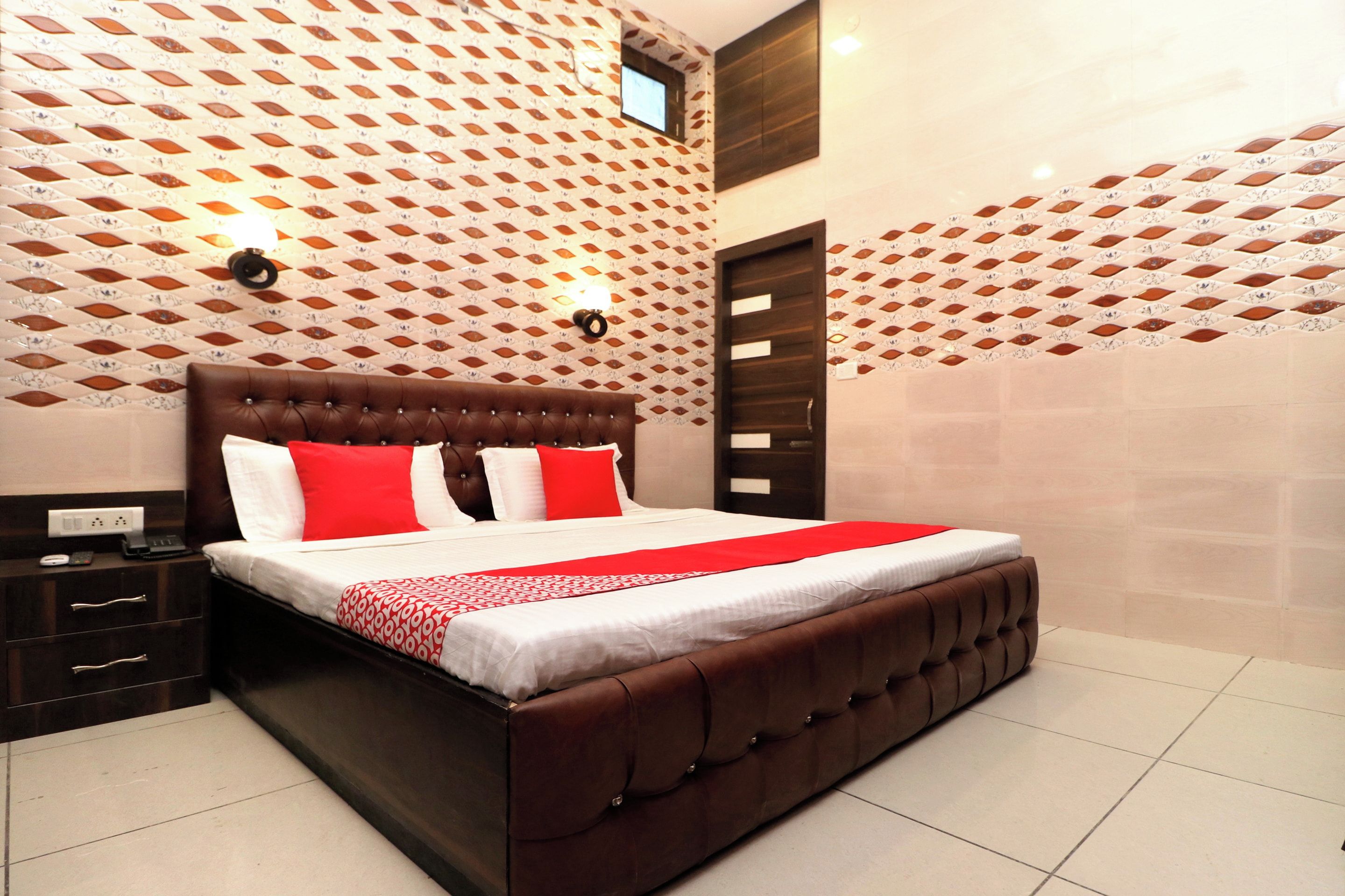A bedroom with a tufted headboard, a bed with white and red bedding, a nightstand with a lamp, and patterned walls.
